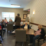 a man standing in front of a group of residents who sit at a common area inside a senior care facility