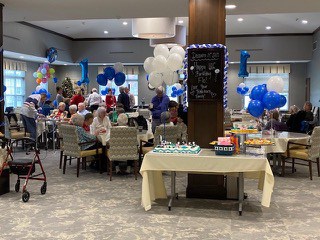 residents of a senior living community enjoying a birthday party with several blue and white balloons attached to tables