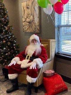 a man dressed as Santa Claus sitting at a chair next to a lit Christmas tree and a candy bowl on top of a stuffed red bag