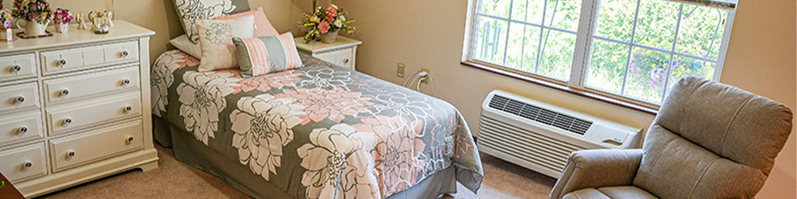 View of a senior living apartment bedroom with a bed that has a pink and grey floral comforter, a grey chair, and a white dresser
