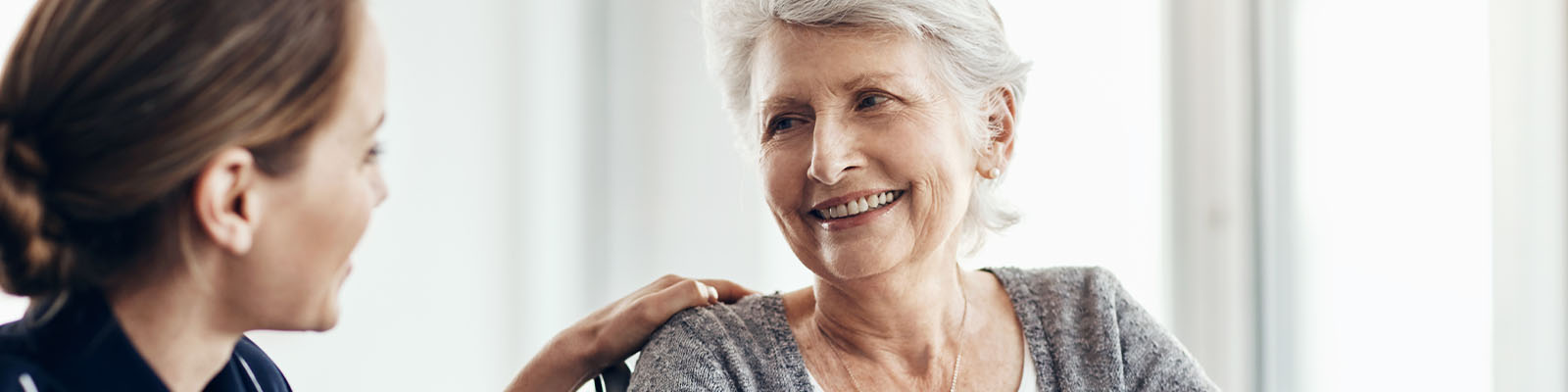 A senior woman in a grey sweater sitting in a wheelchair while looking over at her female nursing assistant in dark blue scrubs