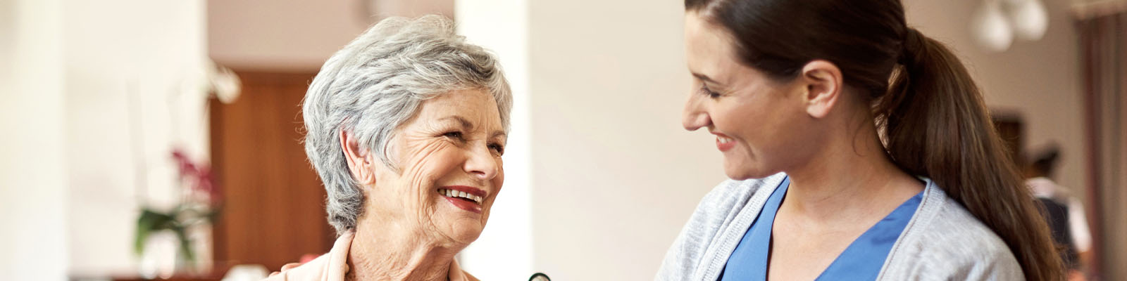 A senior woman sitting in a wheelchair looking over and smiling at her female nursing assistant
