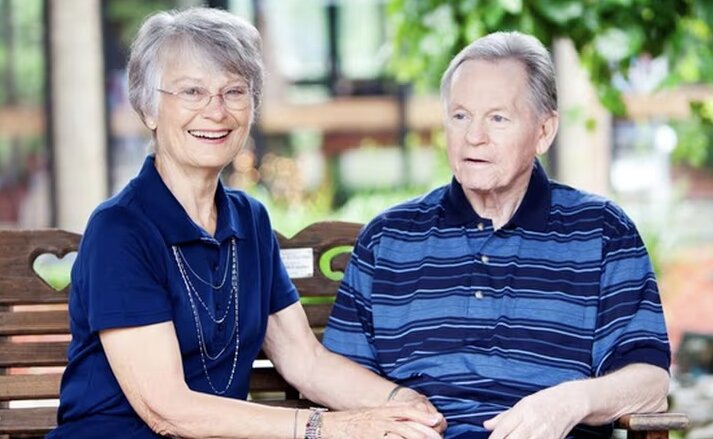 Elderly couple are sitting outside together on a wooden bench. They are wearing matching blue shirts, holding hands, and smiling. Traditions Park Place Senior Living.