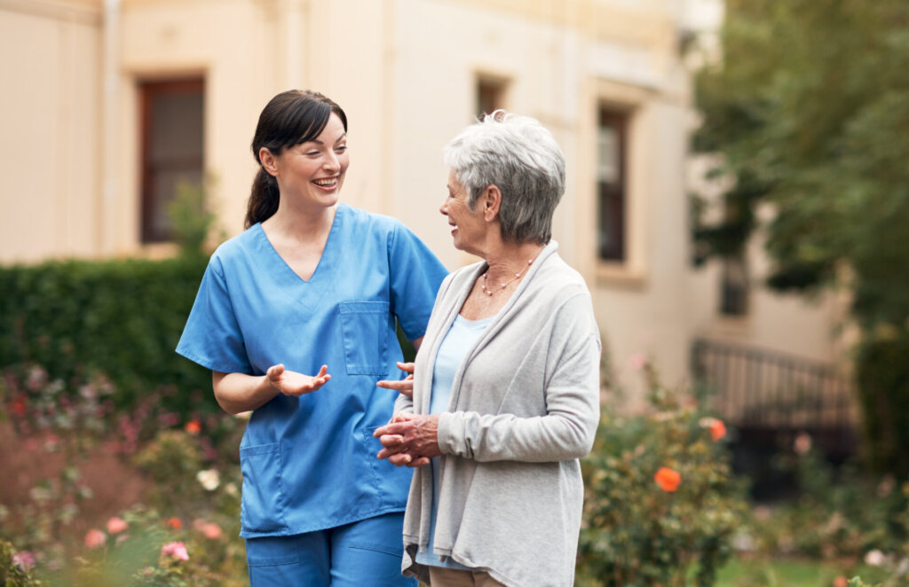A nurse walks with a memory care resident outside. They are having a friendly conversation and enjoying the garden. Traditions Park Place Senior Living.